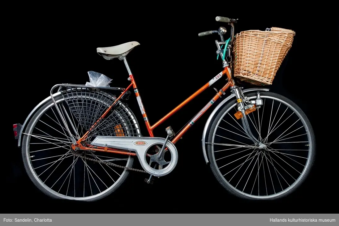 Damcykel i kopparorange färg, metalliclack. Pakethållaren har ett kjolskydd av plast. På styret en cykelkorg. Cykeln är av märket DBS och modellen är Golden Beam. Alla delar till cykeln är original, utom däcken som är utbytta.