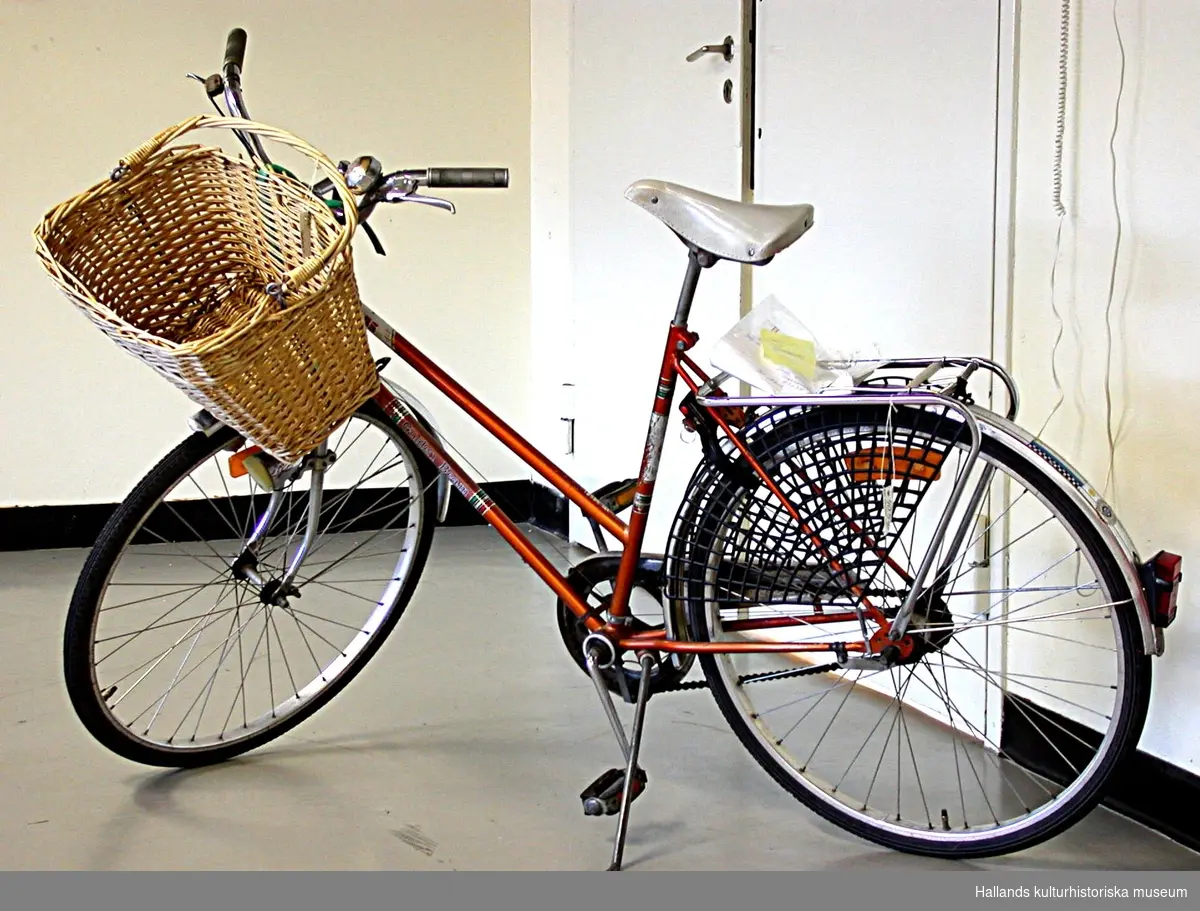 Damcykel i kopparorange färg, metalliclack. Pakethållaren har ett kjolskydd av plast. På styret en cykelkorg. Cykeln är av märket DBS och modellen är Golden Beam. Alla delar till cykeln är original, utom däcken som är utbytta.