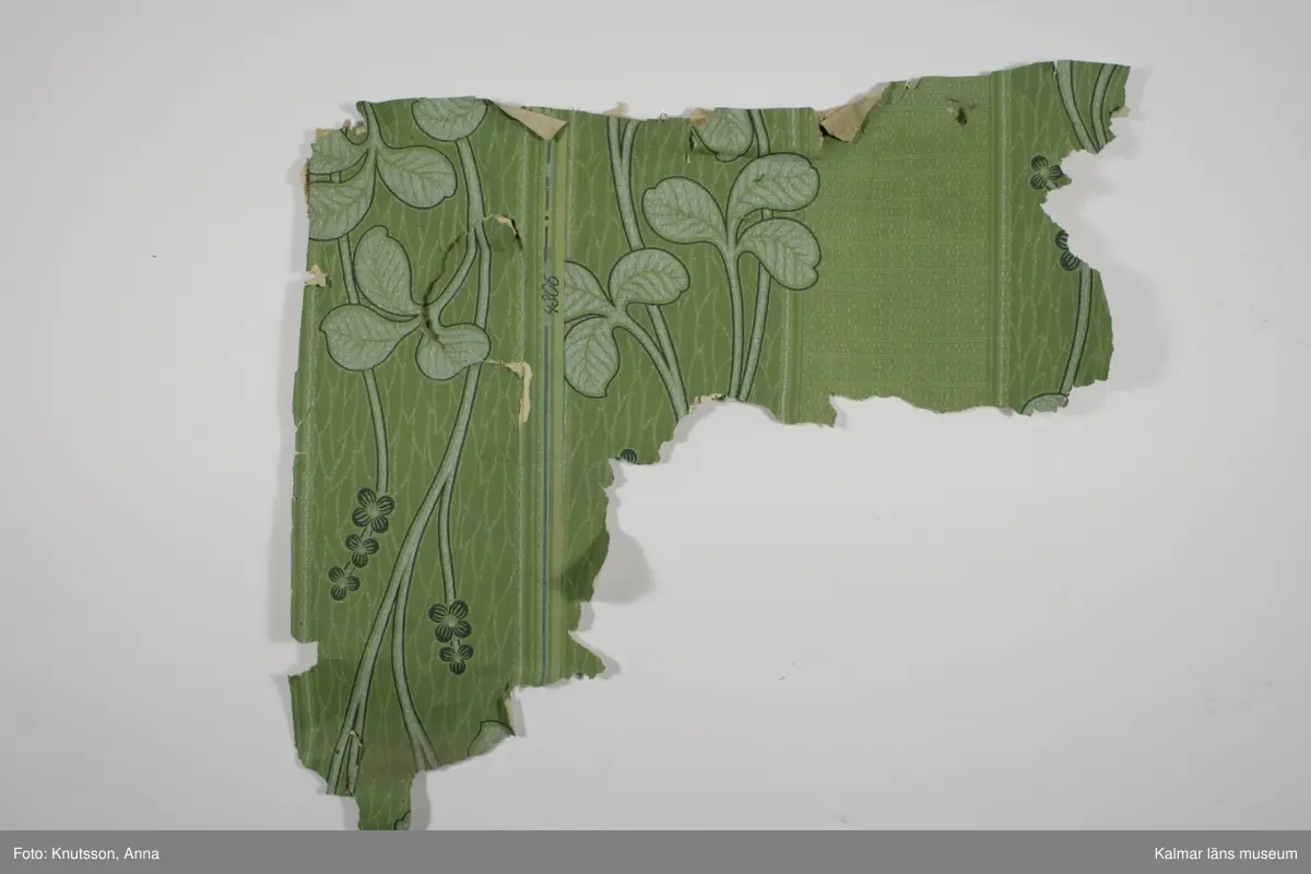 KLM 43950 Tapet i papper. Ljusgrön jugendtapet med bakgrund med lodräta fält. Ena fältet med små prickar och ränder i det gröna. Det andra fältet har spetsiga gröna blad. Mönstret är slingrande bladrankor med stora ljusa gröngrå blad och små mörka blågröna blommor. I kanten på tapeten står det tryckt: 4306. Tidning på baksidan är tryckt 1940.