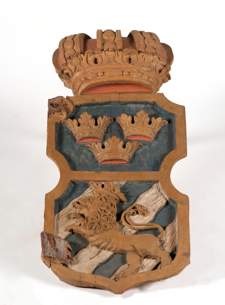 Akterspegelsornament, 2 delar. Dubbelkrönt stora riksvapnet i 2 längsgående hälfter med rester av lejontassar från sköldhållarna. Målad i gult, vitt, och rött på blå botten.