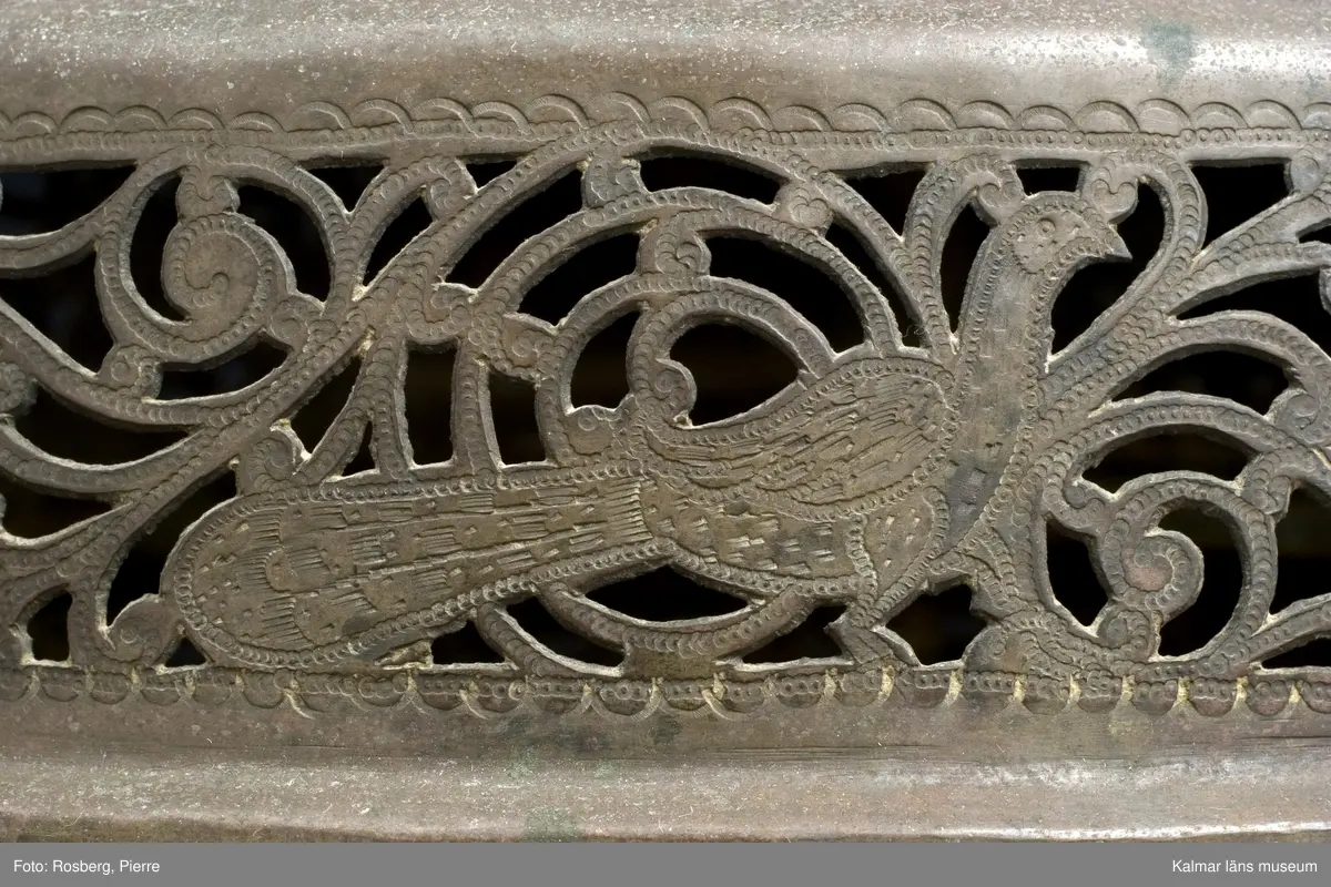 KLM 39725. Lykta. Av mässing. På tre fötter och med ring för upphängning. Med rikt genombrutet rankmönster. Driven mässingsplåt med bucklor på den trappstegsformade överdelen och på sidans nedre del. Tre knoppformiga fötter. I botten drivna bucklor samt en manlig och en kvinlig figur bland slingrande blad och blommor, en heltäckande dekor. Plats för 24 glasrutor, vilka saknas. Inuti tre ljushållare.