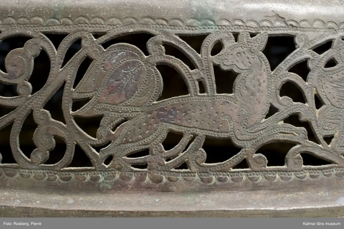 KLM 39725. Lykta. Av mässing. På tre fötter och med ring för upphängning. Med rikt genombrutet rankmönster. Driven mässingsplåt med bucklor på den trappstegsformade överdelen och på sidans nedre del. Tre knoppformiga fötter. I botten drivna bucklor samt en manlig och en kvinlig figur bland slingrande blad och blommor, en heltäckande dekor. Plats för 24 glasrutor, vilka saknas. Inuti tre ljushållare.