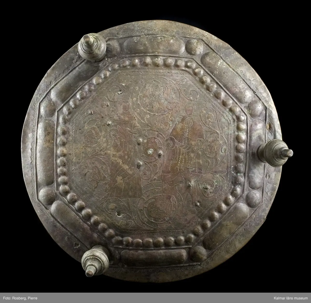 KLM 39725. Lykta. Av mässing. På tre fötter och med ring för upphängning. Med rikt genombrutet rankmönster. Driven mässingsplåt med bucklor på den trappstegsformade överdelen och på sidans nedre del. Tre knoppformiga fötter. I botten drivna bucklor samt en manlig och en kvinlig figur bland slingrande blad och blommor, en heltäckande dekor. Plats för 24 glasrutor, vilka saknas. Inuti tre ljushållare.