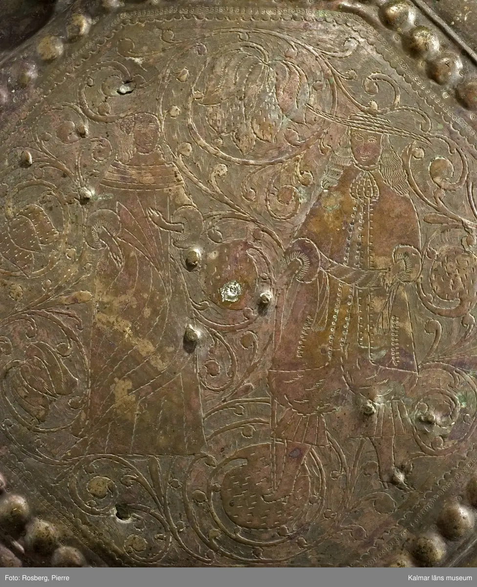 KLM 39725. Lykta. Av mässing. På tre fötter och med ring för upphängning. Med rikt genombrutet rankmönster. Driven mässingsplåt med bucklor på den trappstegsformade överdelen och på sidans nedre del. Tre knoppformiga fötter. I botten drivna bucklor samt en manlig och en kvinlig figur bland slingrande blad och blommor, en heltäckande dekor. Plats för 24 glasrutor, vilka saknas. Inuti tre ljushållare.