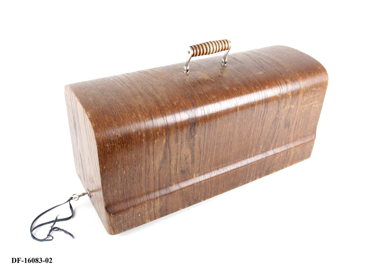 Symaskin på tresokkel, med tilhørende lokk med lås og håndtak. Symaskinen er utstyrt med lampe og  elektrisk motor, festet på baksiden med ledning og pedal.