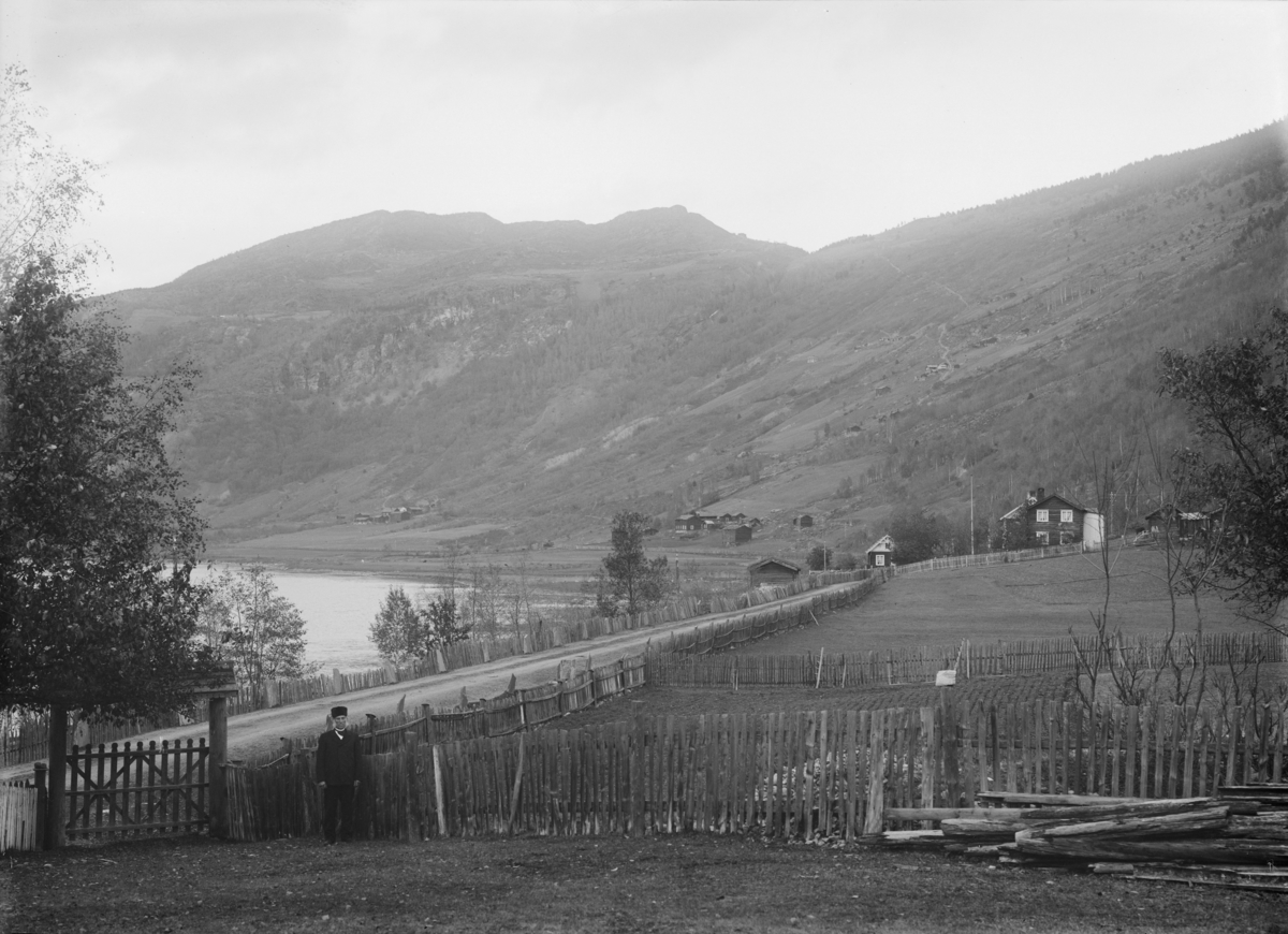 07.10.1909. Parti fra Bredebygden. Bebyggelse, vei, gjerder, mann ved port, åsside med bebyggelse og skog, elv.