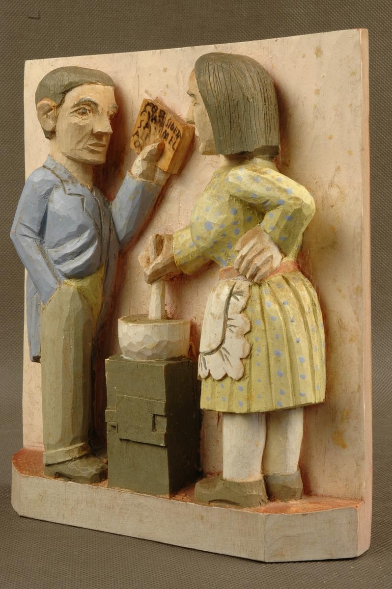 Kvinne- og mannsfigur. Mannen holder en bok, kvinnen rører i en gryte. Motivet inngår i en motivkrets fra hjemmet og det dagligdagse.