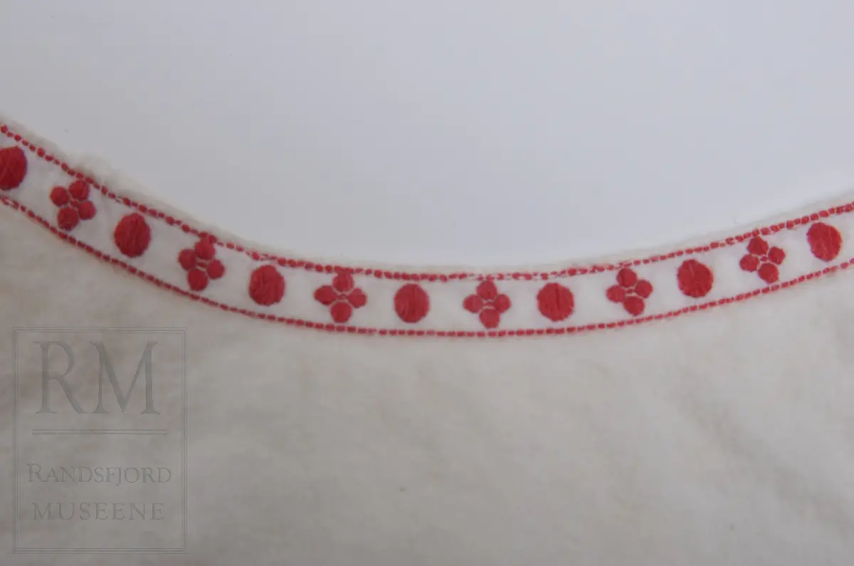 Liv til løyert e.l. til spebarn. Hvit flanell fóret med hvitt kypertvevet bomullstoff. Flanellen har lyse fargestriper langs kantene. Livet er kantet mot bomullsbpnd med rødt mønster. Omslagsliv med bendelbånd til å knytte med. 3 Lerretsknapper.