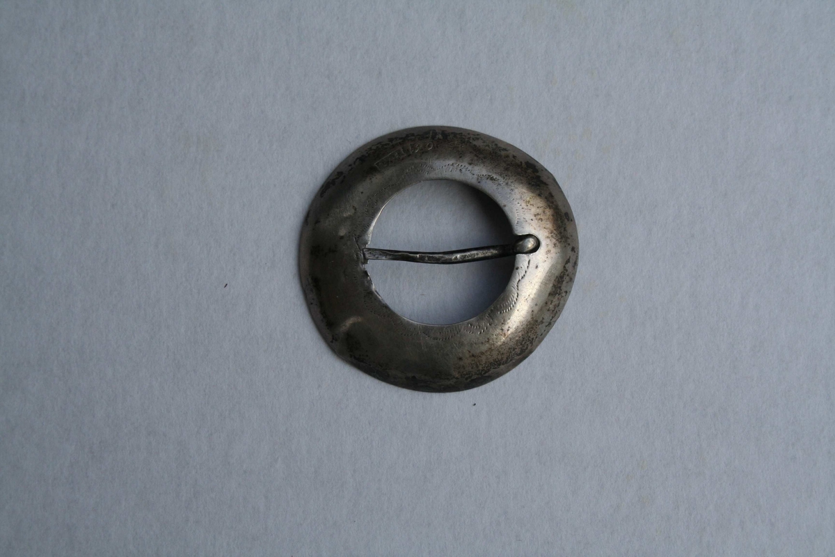 Dette er ein flat ring av sølv, laga av ei sølvplate. Den er litt gravert og har fått ei litt bua form. Ingen lauv. Festes med ei tann gjennom stoffet. 