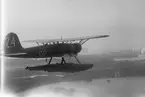Flygplan S 12 märkt nummer 24 tillhörande F 2 Hägernäs i luften över skärgård. Flygplanet sett bakifrån.