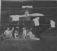 Sommardag. Fem män med bar överkropp sitter framför ett flygplan i en hangar på Hästholmens flygfält, 1923. På flygplanspropellern hänger tröjor.