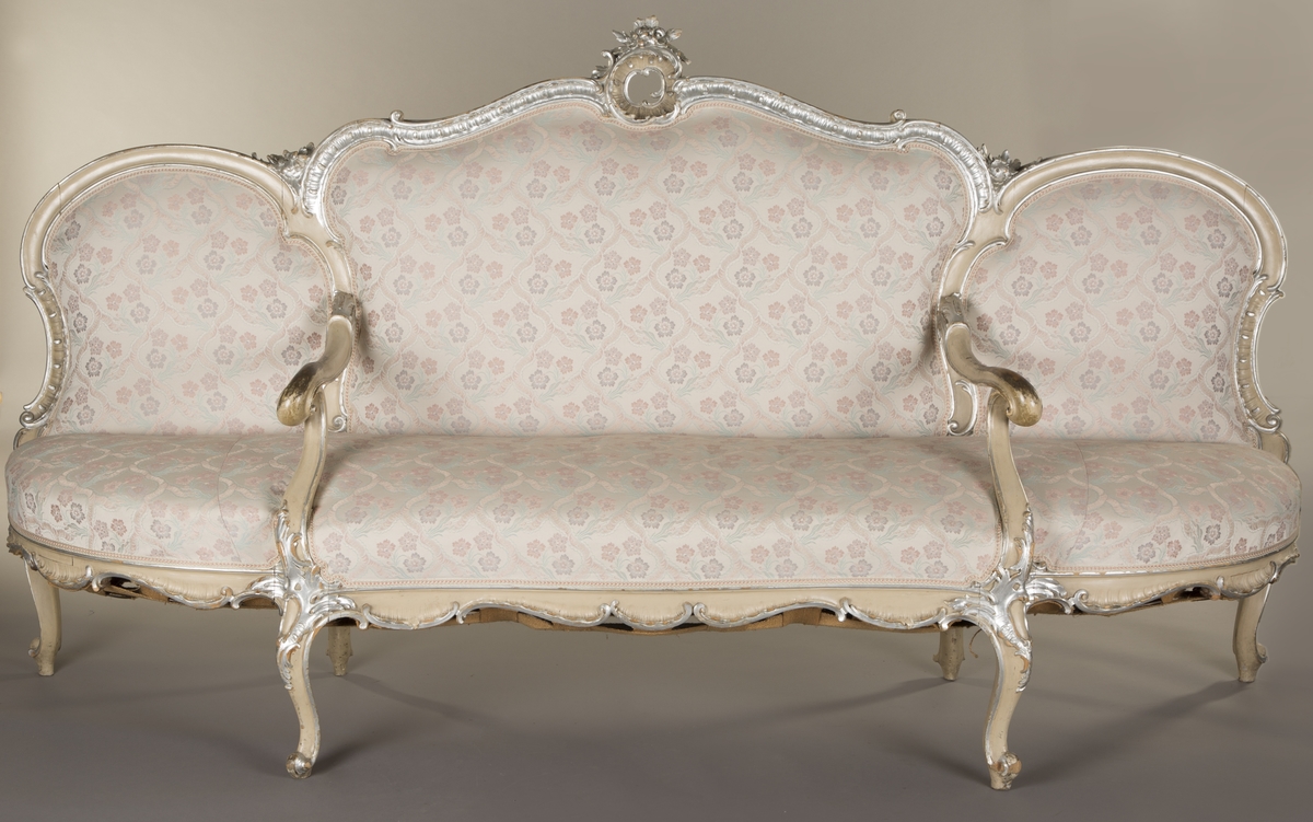 Sittemøbel for flere personer. Nyrokokko. Canapé à confidente - type, d.v.s. på hver side avsluttet av et sidesete. Alt treverk malt lyst grått med forsølvede ornamenter. Høy, stoppet rygg. Spiralfjærer både i sete og rygg. Trukket med tykk, rosa silke med ivevete, flerfargete blomster.
6 cabrioleben. de to forbena har øverst utskåret rocaille - lignende motiv med små blomster. Et glatt, åpent armlene på hver side av midtstykket.