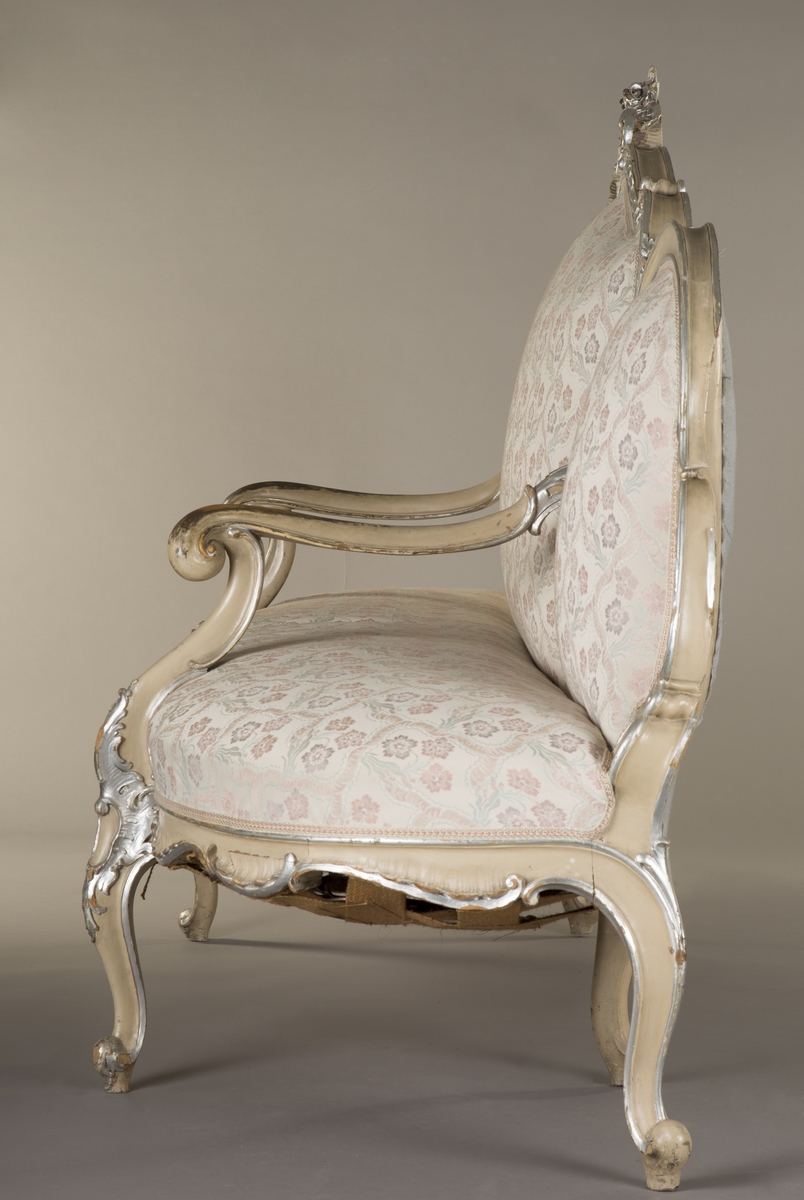 Sittemøbel for flere personer. Nyrokokko. Canapé à confidente - type, d.v.s. på hver side avsluttet av et sidesete. Alt treverk malt lyst grått med forsølvede ornamenter. Høy, stoppet rygg. Spiralfjærer både i sete og rygg. Trukket med tykk, rosa silke med ivevete, flerfargete blomster.
6 cabrioleben. de to forbena har øverst utskåret rocaille - lignende motiv med små blomster. Et glatt, åpent armlene på hver side av midtstykket.