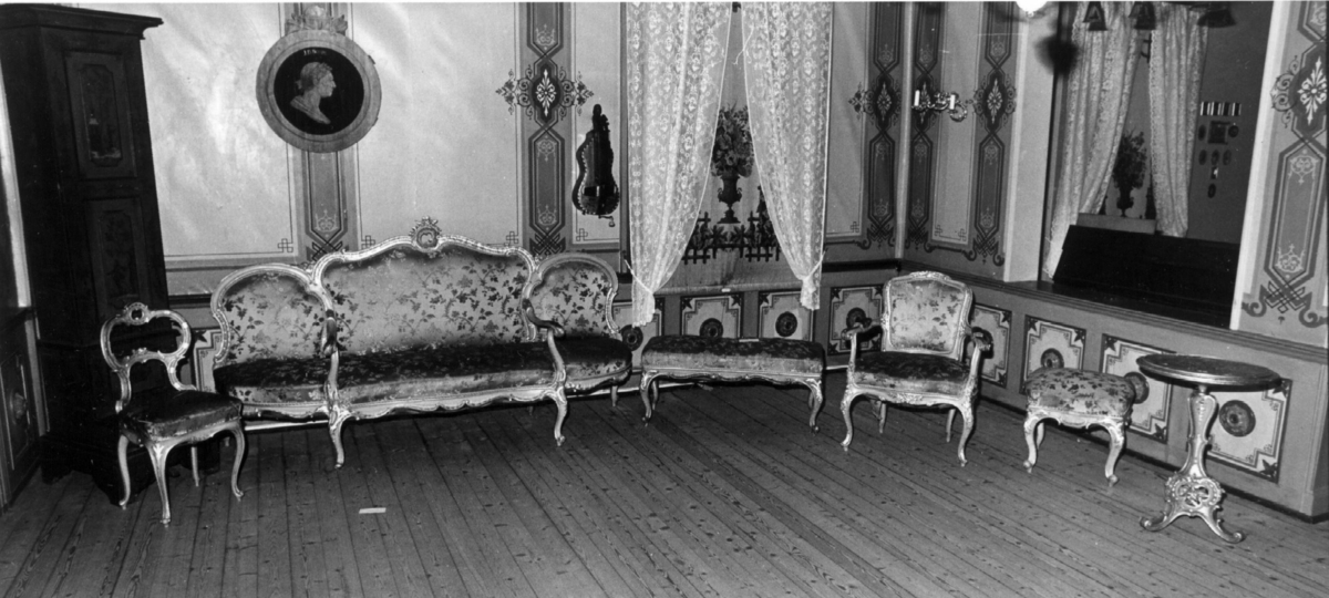 Sittemøbel for flere personer. Nyrokokko. Canapé à confidente - type, d.v.s. på hver side avsluttet av et sidesete. Alt treverk malt lyst grått med forsølvede ornamenter. Høy, stoppet rygg. Spiralfjærer både i sete og rygg. Trukket med tykk, rosa silke med ivevete, flerfargete blomster.
6 cabrioleben. de to forbena har øverst utskåret rocaille - lignende motiv med små blomster. Et glatt, åpent armlene på hver side av midtstykket.