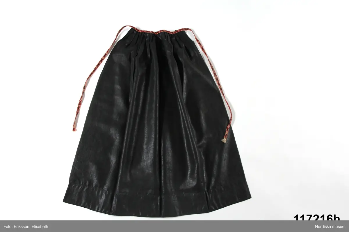 Huvudliggaren:
"Sorgdräkt för kvinna. Särk med 'hängseltåg', 'särkyvdel'; vit -'häsklä'; blått livstycke; svart tröja; huvudduk; 'svart näsduk'; hatt; 'raskförklä', 'sockar',  'hårbandsvifsa', huvudbindel."

Bilaga:
a: "8 Särk, övdel. av linne eller blågarn. med broderade linningar på överdelen"
c: "11. Hvit häsklä."
Anm: Kung Kerstin Larsdotters resa i Floda sn Dalarna 24/2 - 8/3 1912. Innehåller förteckning på inköp av dräktdelar från Floda, inv nr 117 216 - 117 239. Här finns  tillverkningstid och beskrivningar på vissa av dräktdelarna.  I vissa fall nämns också utförligt på vilka helgdagar dräktdelen  användes.

Katalogkort:
a: Hängselsärk
b. Överdel av halvlinne vävt i kypert.
c.  Halskäde av småmönstrat bomullstyg, liknande kambris, vävt  i kypertvariation med drällmönster.
d. Livstycke av mörkblå kraftig vadmal,
e. Tröja av grov svart vadmal

i. 
 Mönstervävt band med upplockat mönster i rött z-tvinnat ullgarn på vit linnebotten, med hjärtan, kryss och rombformer, 2 band om vardera 124 cm har vikts och sytts ihop så att det ena bildar en ögla och det andra har fria ändar avslutade med en tofs i bandets garn.
Läs mer om dräktskicket i Floda i: Albert Alm, Dräktalmanacka för Floda socken, Nordiska museet, Sthlm 1930
/Berit Eldvik 2005-04-14