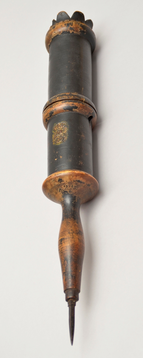 Bodstikke. Uthola sylinder i to delar.
Inni holrommet ligg eit samanrulla ark, papir er fest til eit tynt vove ark. Det er handskrive tekst på arket. Noko av byrjinga er borte, då noko av papiret er falt av der.
Sylindaren er i tre med ein jarnspiss i eine enda. Det er to små, gjennomgåand hol nedst på kvar del. Når dei to delane var sett saman, vart det sett inn ein pinne eller liknande gjennom hola på kvar side, slik at delane vart låste saman.
På eine sida og på enda er det eit symbol stansa inn i treet. Symbolet ser ut som ein stor C med noko inni. Over C-forma er ei krune.