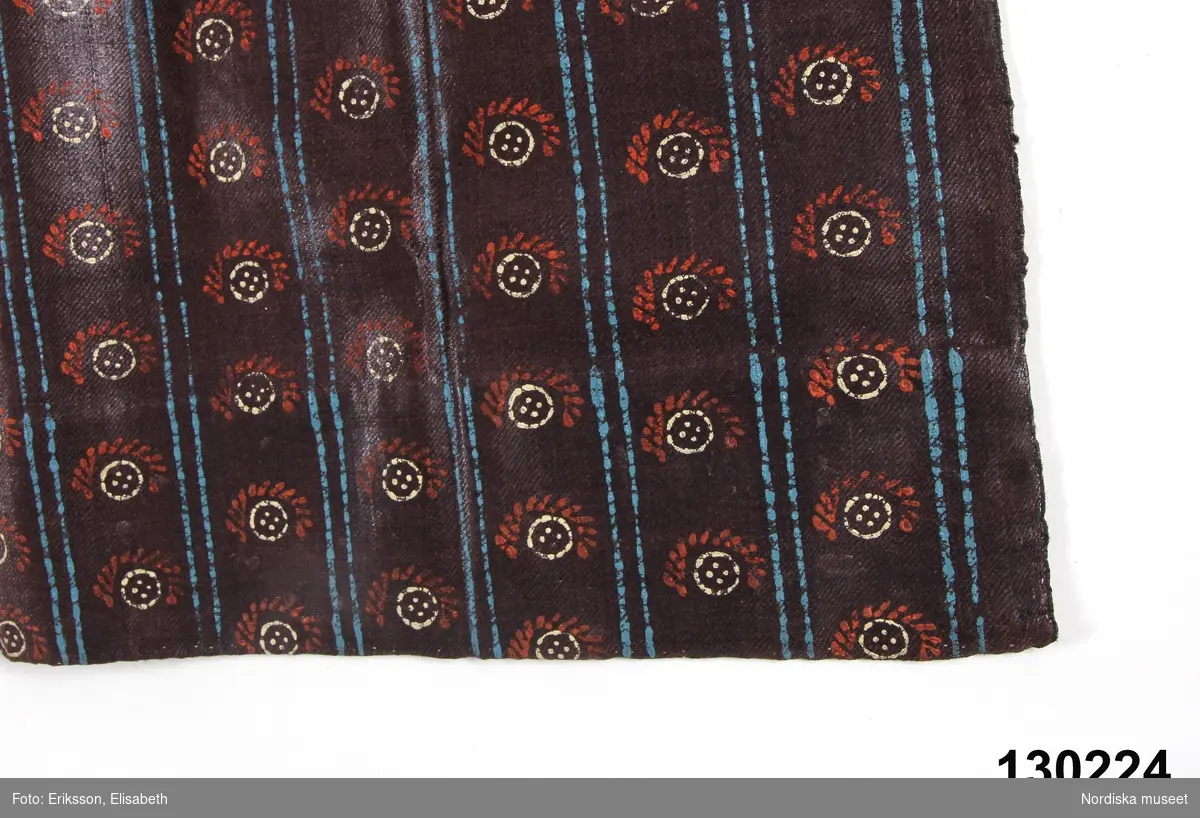 Förkläde av mörklila tunn yllekypert av kamgarn med starkt glättad yta, s.k. rask eller chalon. har fått ett sekundärt stämpeltryckt mönster med oljefärg, smala dubbla  ränder  i klarblått och mellan ränderna rader av  "rosor" dvs. mönsterfigurer i vitt och rött. Rynkat i midjan med ett påsytt  3 cm brett midjeband i ylle smårutigt  i rött, grönt och vitt av den sort som kallas Malsband dvs. Malungsband. Nederkanten inskodd med  smalrandigt halvylletyg i grått och grönt/ljusgult. Handsömnad.
Rask eller chalon var ett köpetyg som användes till högtidsförkläden. Här har man låtit en lokal tryckare mönstra tyget, vilket är ovanligt på denna typ av förkläde. 
erit Eldvik 2012-03-29