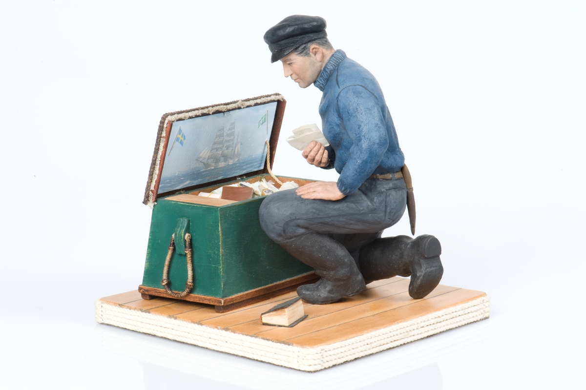 keramikfigur av en sjöman vid sin sjömanskista. Flera små detaljer i kistan. Bl a fartygsporträtt av barkskepp, målat av Hendel Friberg.