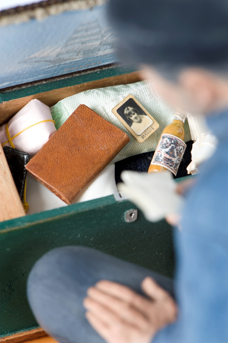 keramikfigur av en sjöman vid sin sjömanskista. Flera små detaljer i kistan. Bl a fartygsporträtt av barkskepp, målat av Hendel Friberg.