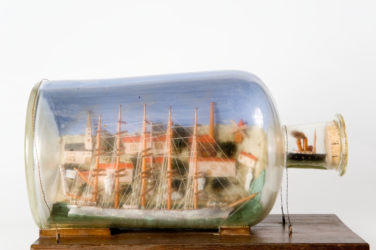 Flaskskepp med femmastad fullriggare. Seglen är beslagna och i flaskans hals är en liten bogserbåt behjälplig. I bakgrunden är en bebyggd strand. Tillverkaren är okänd.