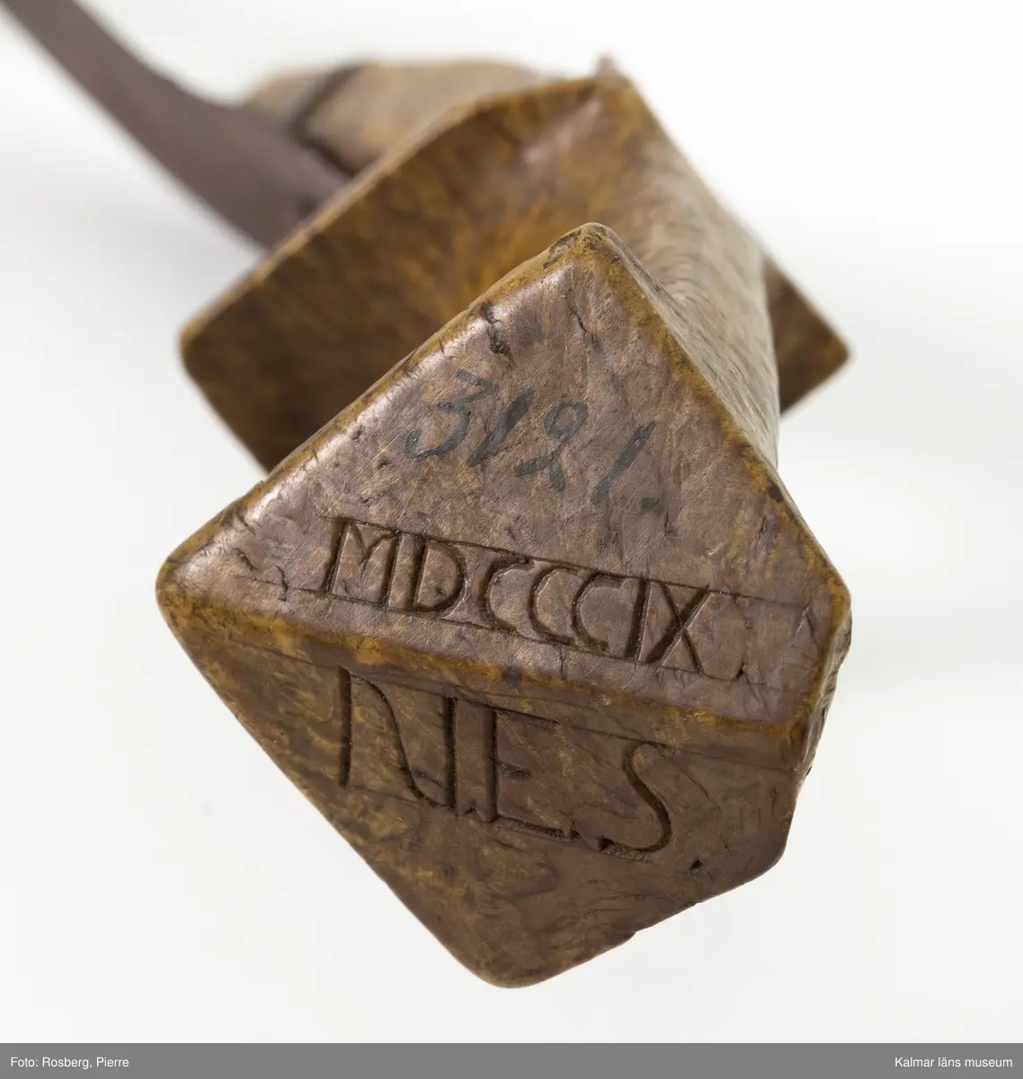 KLM 3121. Skära, vinkelskära, trä, metall. Vinkelskära med bågböjt blad av järn, avbrutet, fastnitat på skaftets ena sida. Skaft av masurbjörk med utskuret handtag och fyrkantig knopp, upphöjd på mitten. Bladet är målat i rödbrun färg under senare tid. På knoppens undersida inskuret monogramet N.E.S. och årtalet MDCCCIX (1809). Nära bladet brännstämplat PNS. Bladet märkt A. Användes till att skära av säden.