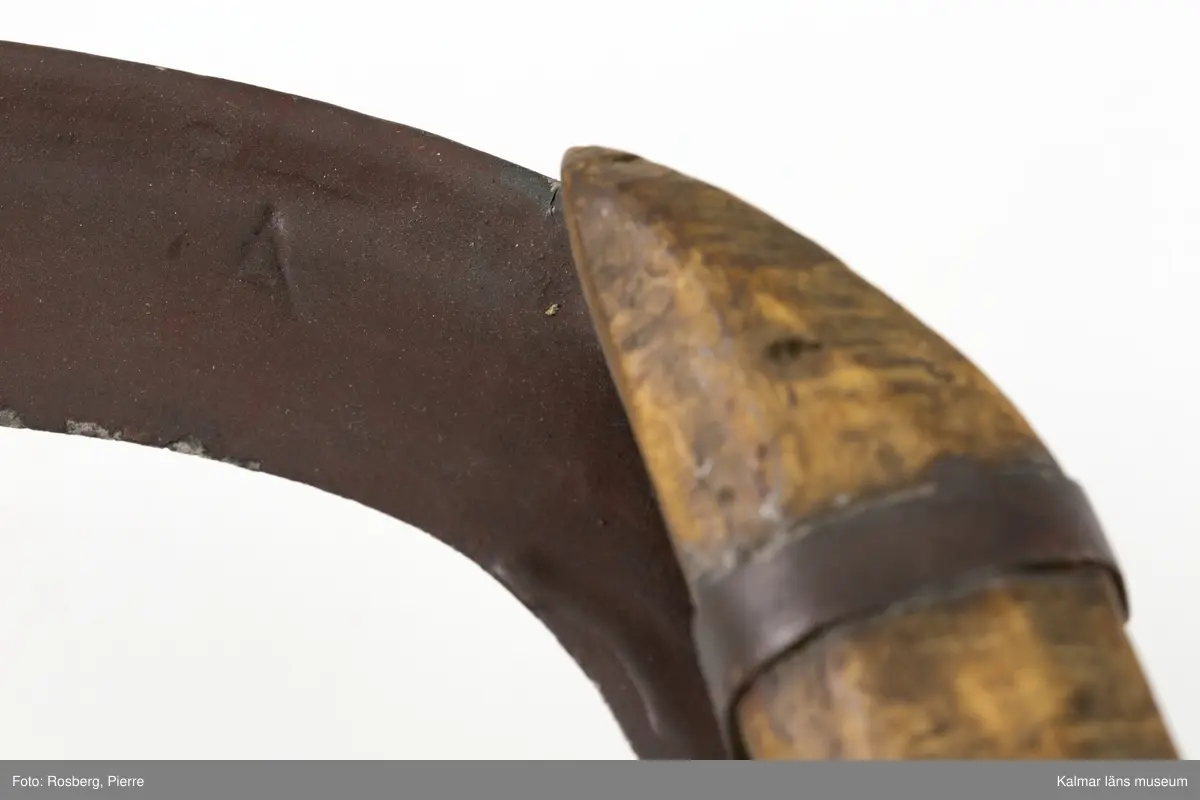 KLM 3121. Skära, vinkelskära, trä, metall. Vinkelskära med bågböjt blad av järn, avbrutet, fastnitat på skaftets ena sida. Skaft av masurbjörk med utskuret handtag och fyrkantig knopp, upphöjd på mitten. Bladet är målat i rödbrun färg under senare tid. På knoppens undersida inskuret monogramet N.E.S. och årtalet MDCCCIX (1809). Nära bladet brännstämplat PNS. Bladet märkt A. Användes till att skära av säden.