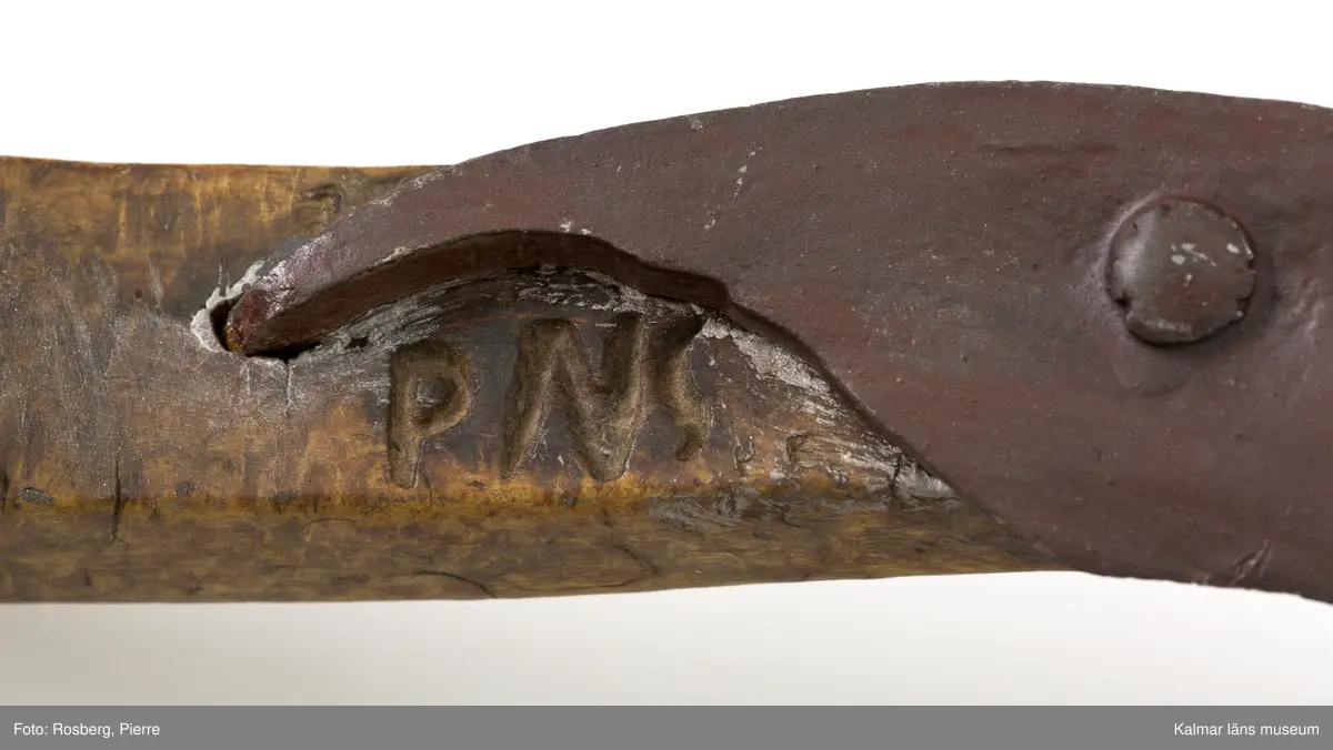KLM 3121. Skära, vinkelskära, trä, metall. Vinkelskära med bågböjt blad av järn, avbrutet, fastnitat på skaftets ena sida. Skaft av masurbjörk med utskuret handtag och fyrkantig knopp, upphöjd på mitten. Bladet är målat i rödbrun färg under senare tid. På knoppens undersida inskuret monogramet N.E.S. och årtalet MDCCCIX (1809). Nära bladet brännstämplat PNS. Bladet märkt A. Användes till att skära av säden.