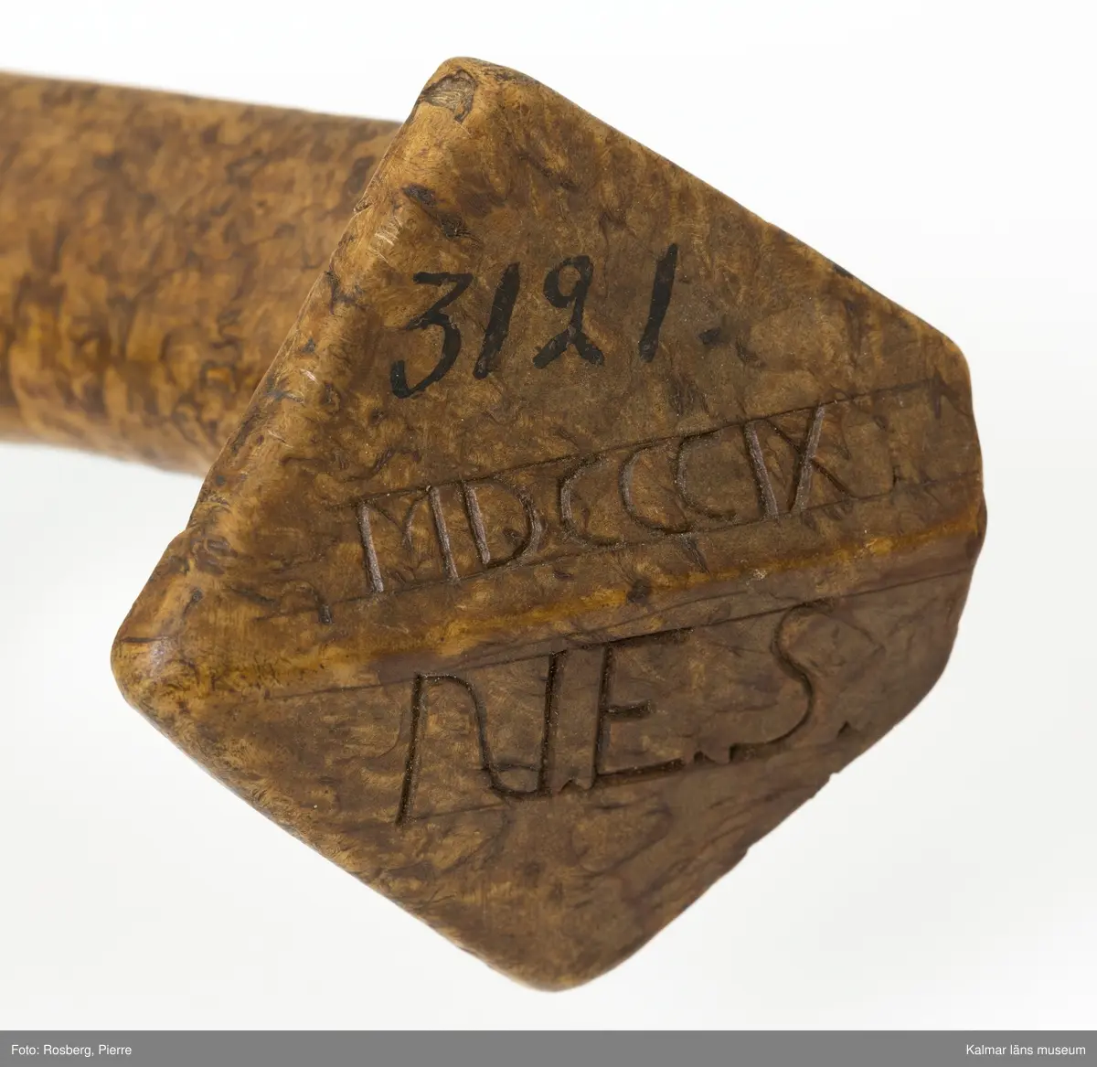 KLM 3121. Skära, vinkelskära, trä, metall. Vinkelskära med bågböjt blad av järn, avbrutet, fastnitat på skaftets ena sida. Skaft av masurbjörk med utskuret handtag och fyrkantig knopp, upphöjd på mitten. Bladet är målat i rödbrun färg under senare tid. På knoppens undersida inskuret monogramet N.E.S. och årtalet MDCCCIX (1809). Nära bladet brännstämplat PNS. Bladet märkt A. Användes till att skära av säden.