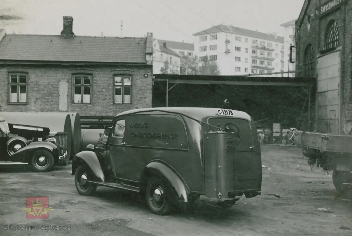 "Karbidgenerator produsert av Øveråsens Motorfabrikk, Gjøvik. De ble montert av Colbjørnsens & Co. A/S under hele krigstiden." I følge merking i album. På bilen med kjennetegn C-12178 står det skrevet "A/S Norske Konfektionsfabriker." På skiltet øverst på murbygningen står sannsynligvis "A/S Sveiseindustri." 

Bilde 1/4 er en Chevrolet med en eldre Chev. baklykt  på taket. 20.08.2016: "Bilde 1 er en Chevrolet 1939-40 varebil med norskbygd karosseri. "25.06.2018: Til venstre snuten av en Packard fra sent 1930-tall. Opplysninger fra Ivar Erlend Stav.
Klikk på pilen til høyre:
Bilde 2/4 er en Dodge 1935-1936 modell. (Strømmenbygd?)
Bilde 3/4  er en 1930 modell Brockway. 25.06.2018: Bildet 3 viser snuten av en Plymouth1937. Opplysninger fra Ivar Erlend Stav.
Bilde 4/4 er en Ford fra 1937-1938 modell." 25.06.2018:  "Ford 1938, siden det var første år med langsgående blanklist hos Ford. Karosseriet er originalt amerikansk varebilkarosseri på personbilchassis ("sedan delivery" - i motsetning til et mer grovbygd varekarosseri på lett lastebilchassis og lastebilfront, kalt "panel delivery". Opplysninger fra Ivar Erlend Stav. 

17.08.2016: Opplysninger til kjøretøyene på bildet v/tidligere daglig leder John Hindklev ved Norsk Kjøretøyhistorisk Museum. 

"Under andre verdenskrig besto virksomheten til firmaet Colbjørnsen stort sett av å montere og vedlikeholde karbidgeneratorer fra Øveråsen. Legg merke til hvordan man felte generatorene inn i varebilkarosseriene. Reklameteksten  ble delvis borte." Sitat fra boka: "Familiefirmaet-utenom allfarvei/2000 årsboken." Colbjørnsen & Co A/S, av Bjørn Ausjen Johannessen.