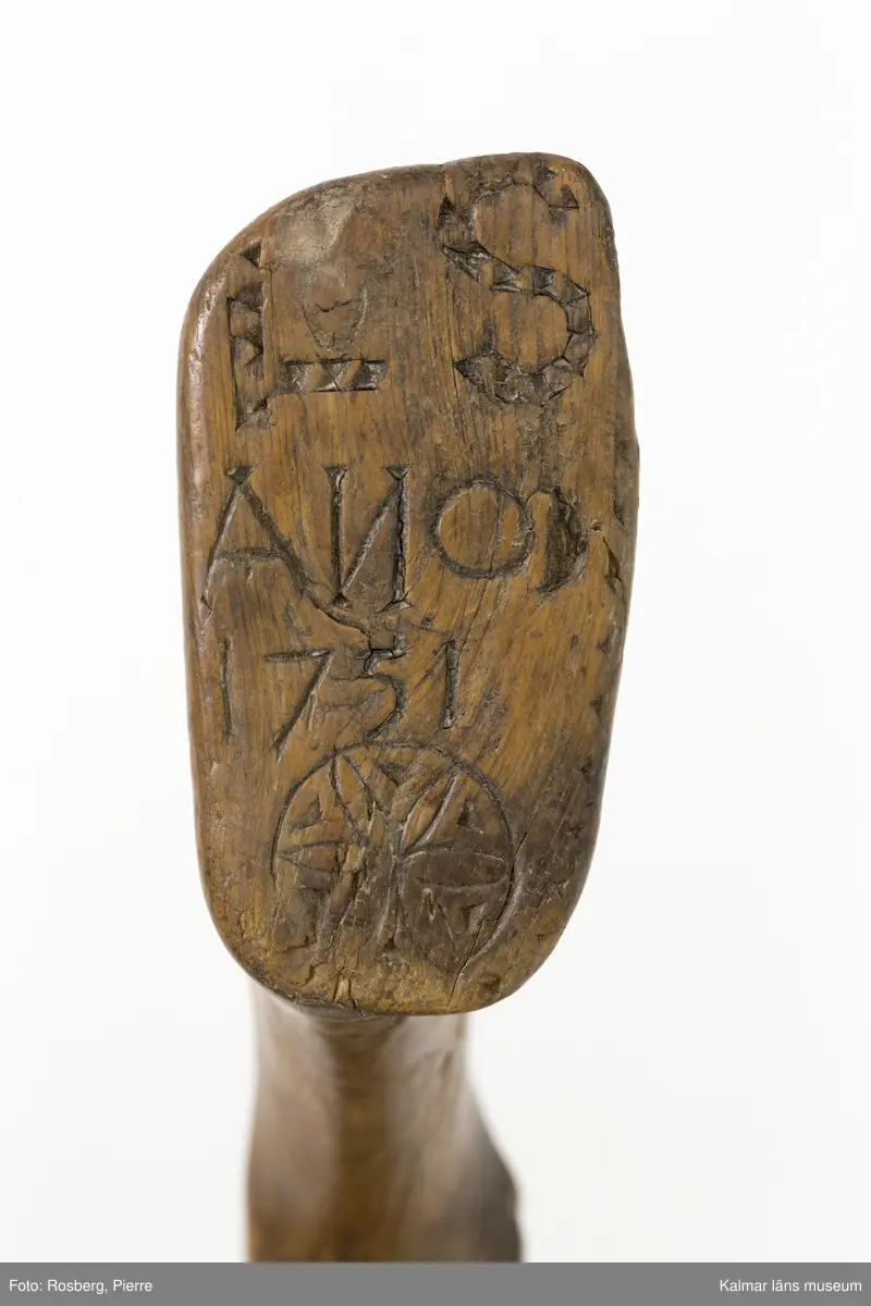 KLM 22639. Skära, vinkelskära. Av trä och metall. Vinkelskära med bågböjt blad av järn, fastnitat på skaftets ena sida. Skaft av trä med utskuret handtag och ovalformad platta i änden. Handtaget har skuren dekor i form av sicksack-mönster. Under skaftet en blomma med ring runt samt "LS" (= Lisa Svensson) ANO 1751. Bladet är målat i rödbrun färg, för rostskydd, under senare tid.