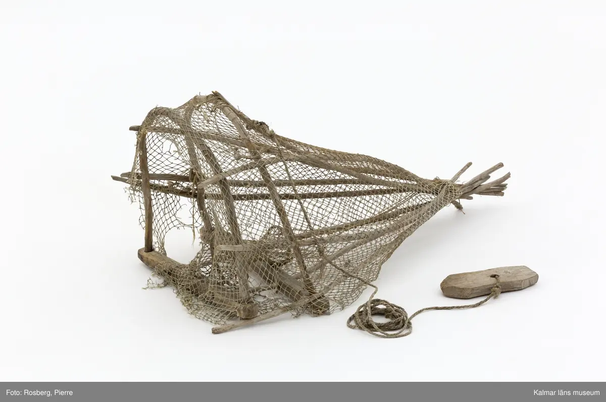 KLM 45863. Mjärde. Avlång fångstbur med stomme av trä och vidjor böjda till bågar med bundet nät av naturmaterial runt om. Även längsgående vidjor som i änden är hopknutna. Inuti en strutformad inredning med en ingång för fisken. På ovansidan ett fastbundet rep med en fastknuten träbit som fungerade som flöte.