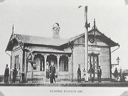 Veddige järnvägsstation som ny år 1880 utmed Varberg-Borås Järnväg, invigd samma år. Stationen är rikt utsmyckad med figursågade dekorer i tidstypisk eklektisk stil, dvs hämtar inspiration från och sammanför element från olika arkitekturstilar. Flera personer står på perrongen varav flera, kanske samtliga, förmodligen arbetar vid järnvägen och stationen.