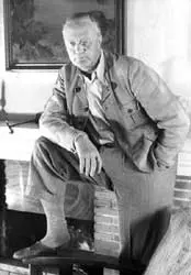 Fotografi av Sigurd Hoel. Han står foran peisen med det ene beinet opp på et armlene. Fotografiet er tatt i svart hvitt. Sigurd Hoel er en eldre herre da bildet ble tatt.