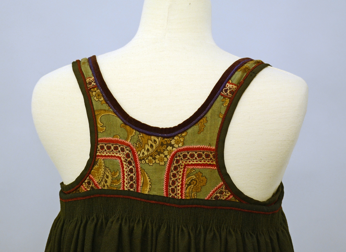 Stakk med påsydd liv. Skjørtet er rynket oppe, metallhekter på hver side av splitten/åpningen foran. Stoffet er av svart vadmel i lerretsbinding. Brodert mørkerød kant øverst på stakken. Skoningen er påsydd med et annet tøy enn det originale, den er 10 cm og har striper av mørkegrønn og sort fløyel.Skoningen er 10 cm. Nest nederste kant er mørkerød.Livet har kjøpetøy i trykteblomster med påsydde bånd. Halsutringningen er av mørkerødt fløyel med broderibord i lilla. Ermutringningene er av sort bomullstøy med mørkerød broderibord. Fòret er av rutet bomullstøy. Stakketøyet er falmet, har noen flekker,og små hull etter møll.