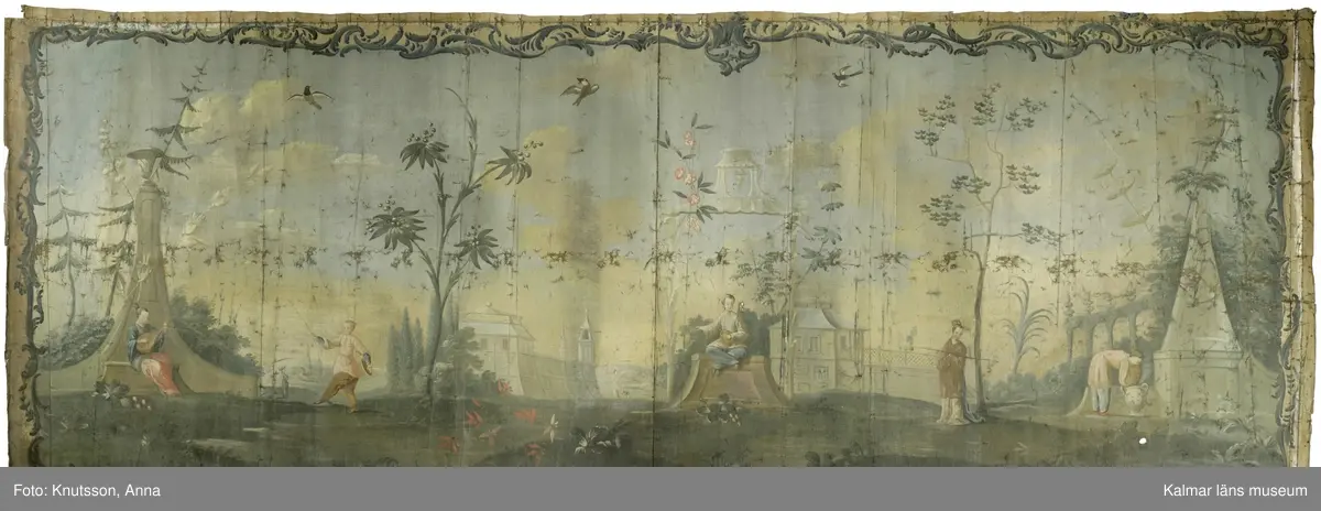 KLM 43973. Tapet, väggfält. Oljemålning på väv. Blå färg utanför ramen. Ramen är snirkliga rankor i mörkblått. Orientaliskt motiv med fem personer i orientaliska kläder. Första personen är en kvinna som spelar ett lutaliknande instrument. Kvinnan har blå överdel och byxor i en lysande röd färg. Nästa person har en ljusgul överdel och beige byxor. Det ser ut som om personen har en flygande fågel fastknuten i ett snöre. I den andra handen håller personen en slags platta. Nästa person sitter ner och håller en likadan platta. Längst till höger i bild står en kvinna böjd över en urna. Kvinnan har rosa överdel, blå byxor och lysande röda skor. I omgivningarna finns flera byggnader, tunna träd, växter och även blommor.