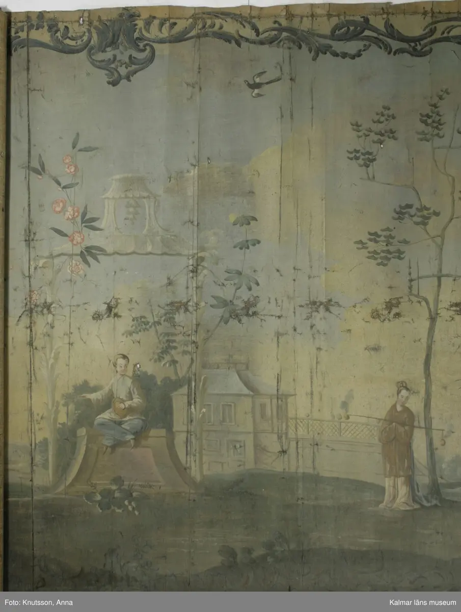 KLM 43973. Tapet, väggfält. Oljemålning på väv. Blå färg utanför ramen. Ramen är snirkliga rankor i mörkblått. Orientaliskt motiv med fem personer i orientaliska kläder. Första personen är en kvinna som spelar ett lutaliknande instrument. Kvinnan har blå överdel och byxor i en lysande röd färg. Nästa person har en ljusgul överdel och beige byxor. Det ser ut som om personen har en flygande fågel fastknuten i ett snöre. I den andra handen håller personen en slags platta. Nästa person sitter ner och håller en likadan platta. Längst till höger i bild står en kvinna böjd över en urna. Kvinnan har rosa överdel, blå byxor och lysande röda skor. I omgivningarna finns flera byggnader, tunna träd, växter och även blommor.