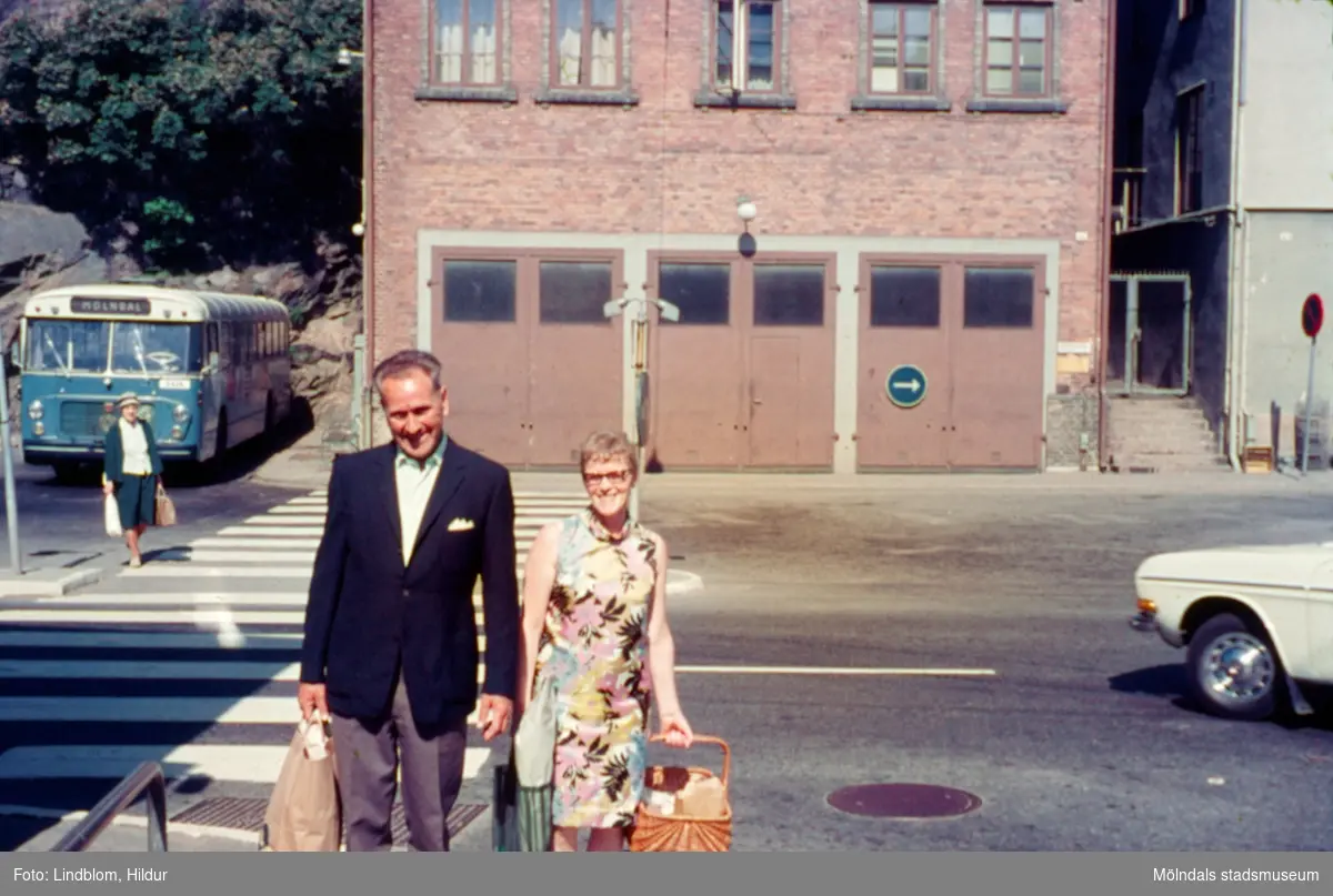 En kvinna och en man vid Gamla Torget i Mölndal, 1970-tal. I bakgrunden till vänster ses en blå buss parkerad på busshållplatsen, i mitten ses huset Kvarnbygatan 41, tidigare brandstation och garage för polisbilar, numera Mölndals målarskola. Till höger ses liten del av huset Kvarnbygatan 43, tidigare Mölndals stadshus, sedermera musikskola.

För mer information om bilden se under tilläggsinformation.