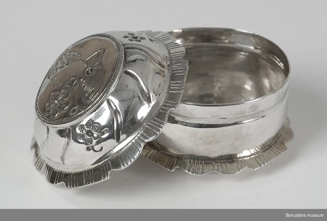 Silverdosa av västsvensk typ i rokokomodell. Rund dosa med godronnerad bård på locket med tre ciselerade blommor. Passformade kanter på lock och underdel med streckutformad godronnering. På locket ciselerad dekor föreställande en kvinna i profil som luktar på en blomma. På underdelens utsida stämplad med Sven Nordströms mästarstämpel SNS, kattfot och stadsstämpel. Årsstämpel saknas. Punktgraverat ägarmonogram i botten på dosan, "B O D". Gravyren möjligen samtida med själva dosan.