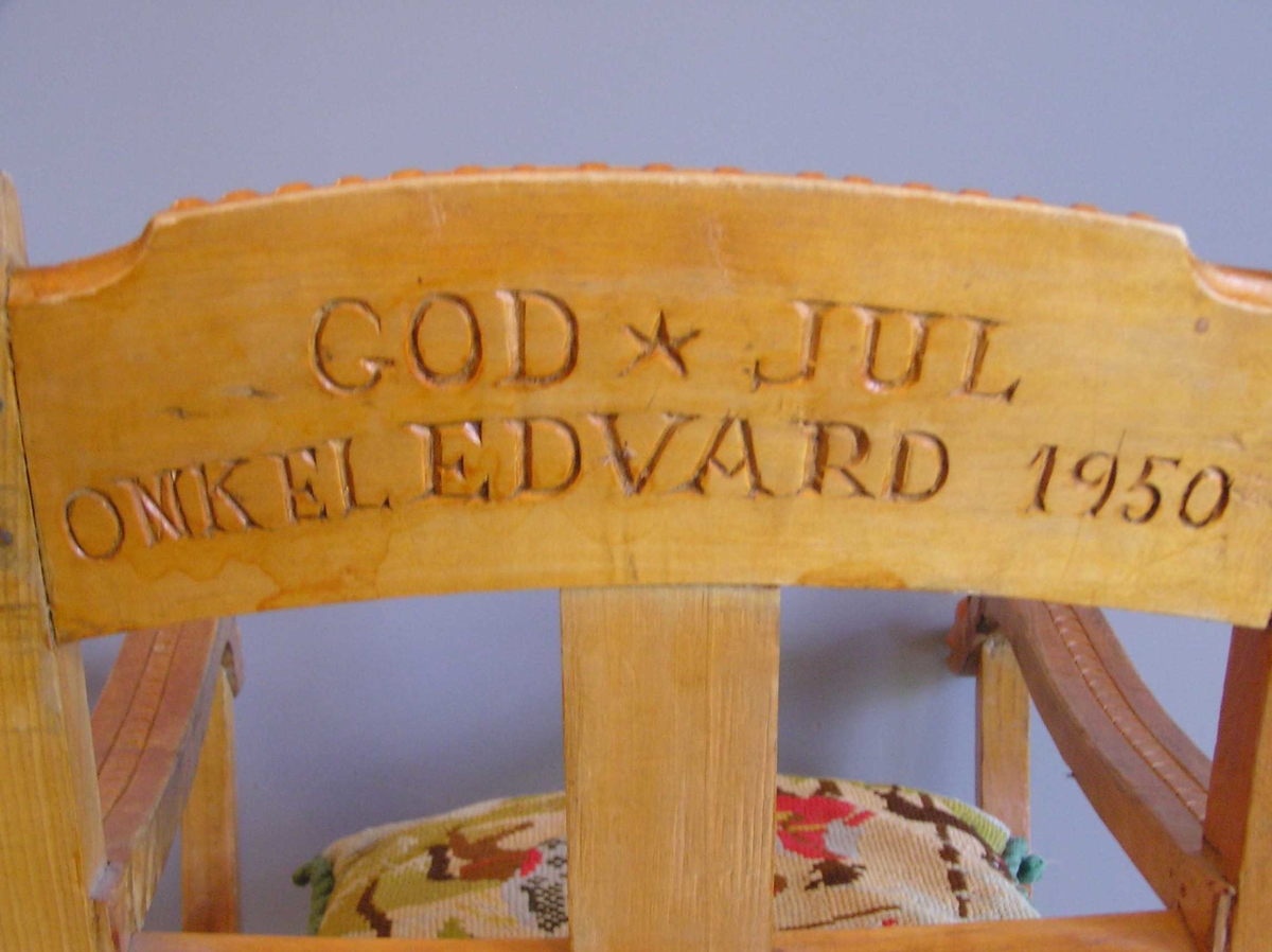 Barnegyngestol med løst sete og løs pute festet med lokker til rygg. Utskåret med figurer.