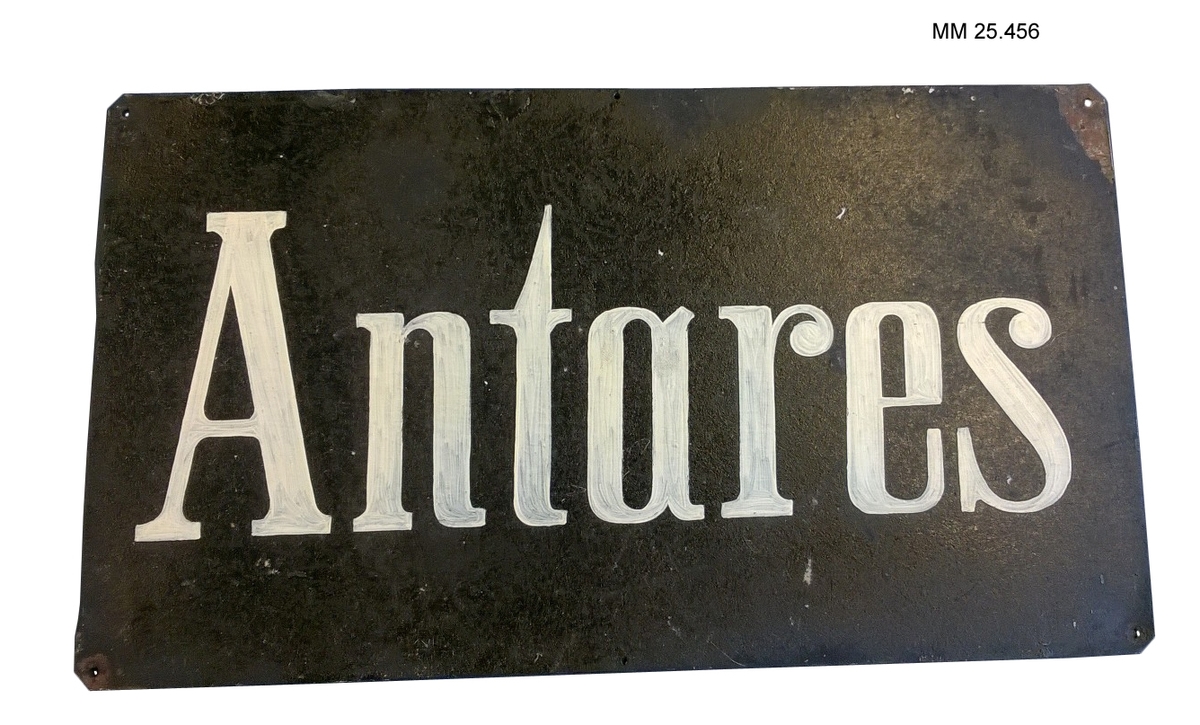 Svart skylt av plåt. Små skruvhål i vart och ett av de fyra hörnen. Text på skylten målad med vit färg där penseldragen syns: "Antares"