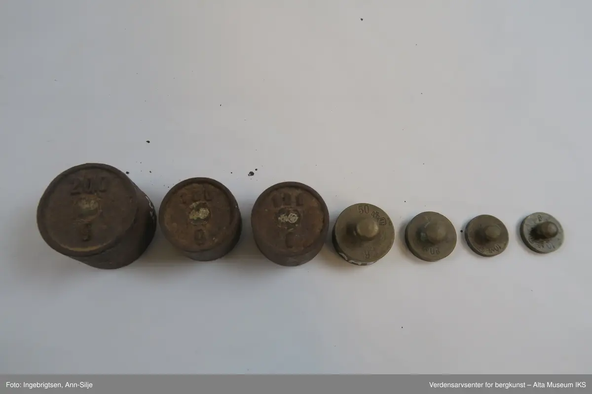 Rektangulær trebit, med hull tilpasset ulike loddstørrelser. Det er 7 lodd: 2 stk. 10 gram, 1 stk. 20 gram, 1 st. 50 gram, 2 stk. 100 gram og 1 stk. 200 gram. Alle loddene er sylinderformet med knott på toppen. Loddene varierer i størrelse: de minste er 2 cm i diameter og 1 cm i høyde, mens den største er 4 cm i diameter og 2,8 cm i høyde.