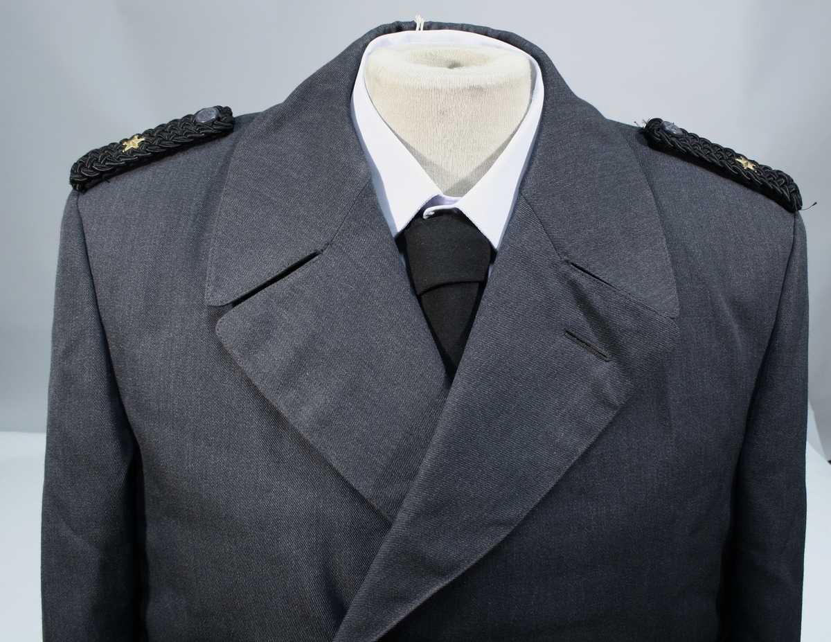 Lang grå uniformsfrakk, grå uniformsjakke, grå vinterlue (båtlue) med for i fuskepels, sort slips, grå uniformslue med blank sort skjerm og regntrekk, tre lyseblå uniformsskjorter. Uniformen har skulderklaffer med en gullstjerne, altså distinksjon for førstebetjent. Plasknapper med valknuten. Luene er forsynt med metallmerker i gull og rødt, den norske krone over valknuten som var symbolet for kriminalomsorgen i perioden 1964-2001.