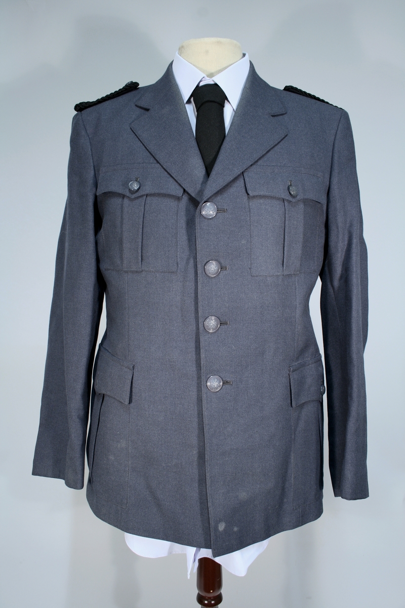 Lang grå uniformsfrakk, grå uniformsjakke, grå vinterlue (båtlue) med for i fuskepels, sort slips, grå uniformslue med blank sort skjerm og regntrekk, tre lyseblå uniformsskjorter. Uniformen har skulderklaffer med en gullstjerne, altså distinksjon for førstebetjent. Plasknapper med valknuten. Luene er forsynt med metallmerker i gull og rødt, den norske krone over valknuten som var symbolet for kriminalomsorgen i perioden 1964-2001.