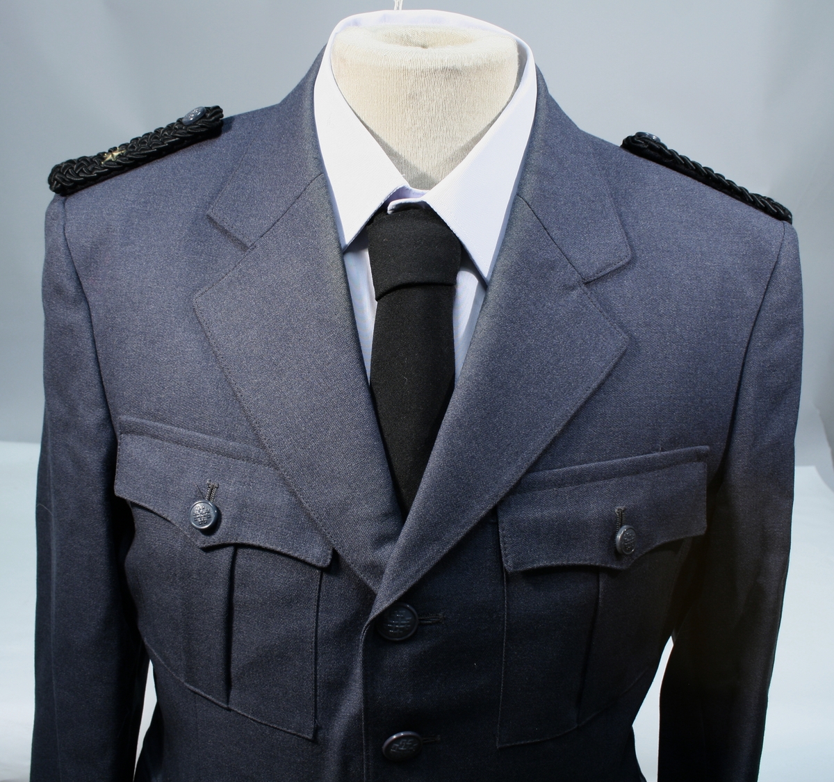 Lang grå uniformsfrakk, grå uniformsjakke, grå vinterlue (båtlue) med for i fuskepels, sort slips, grå uniformslue med blank sort skjerm og regntrekk, tre lyseblå uniformsskjorter. Uniformen har skulderklaffer med en gullstjerne, altså distinksjon for førstebetjent. Plasknapper med valknuten. Luene er forsynt med metallmerker i gull og rødt, den norske krone over valknuten som var symbolet for kriminalomsorgen i perioden 1964-2001.