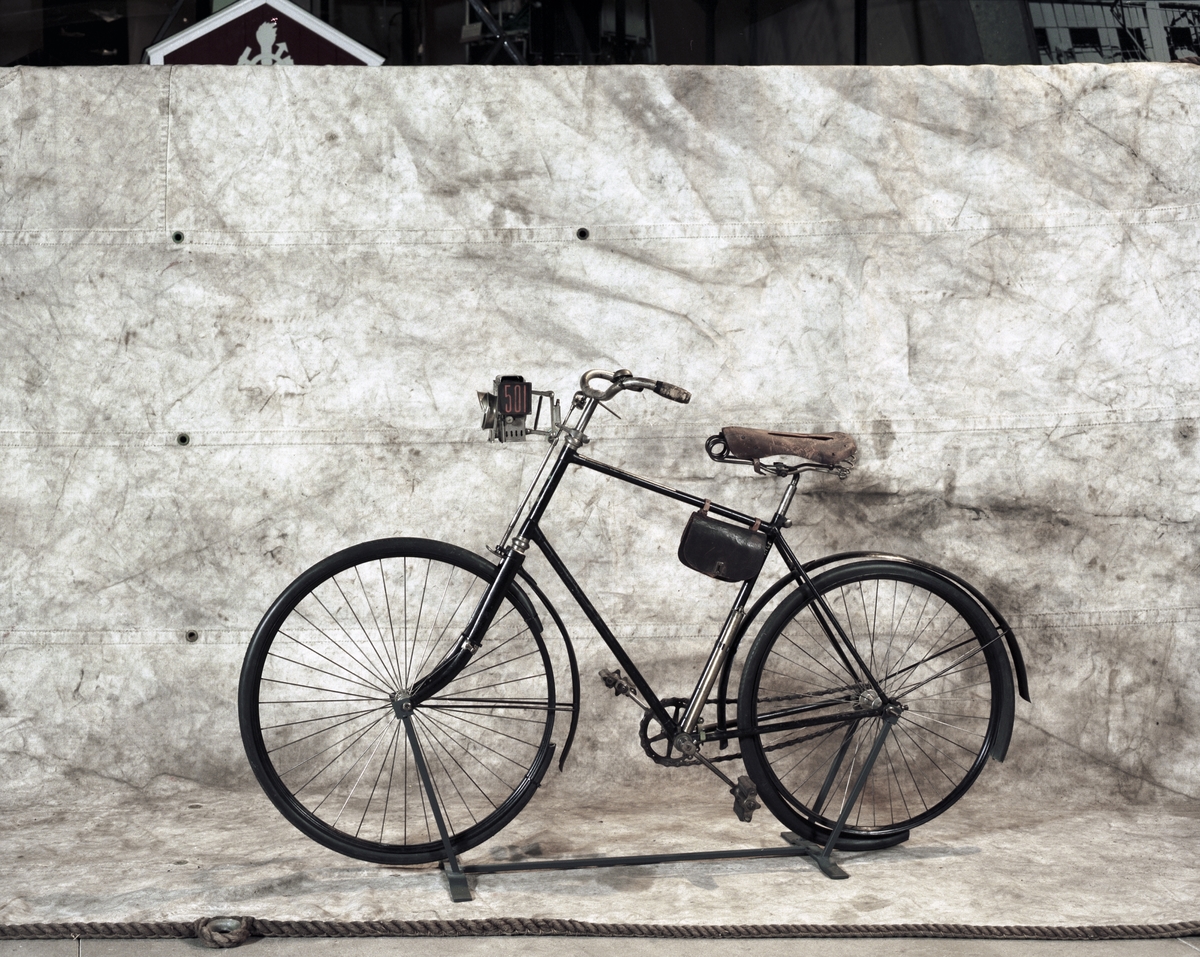 Cykel, "landsvägsmaskin" med ram av stålrör och trådkedjehjul med pneumatiska ringar.
Radiella ekrar. Inget frinav, "gubbpinne" på baknavet för påstigning, fotstöd på framgaffeln att sätta fötterna på vid nedförsåkning.
Skedbroms på framhjulet. Blockkedja, avtagbara stänkskärmar av plåt. Kullagrade nav och vevparti. Ramen är svartlackerad, blanka delar förnicklade. Pris med Dunlop luftringar 1894 kr. 400 kr.
Framhjulet 75 cm i diam, bakhjulet 70 cm.
Lykta: Rovoljelykta med sidoglas, varpå är målat "501" i rött. "Wm Brown's, Rob Roy, Safety lamps". Verktygsväska, vid inköpet tom. Pump för ringarna.