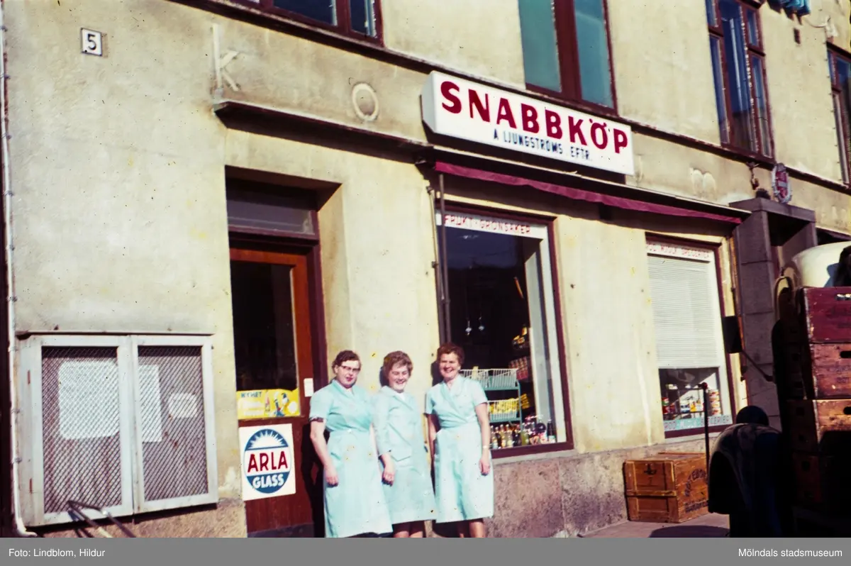 Tre kvinnliga butiksbiträden fotograferade utanför snabbköp med adress Kvarnbygatan 43 vid Gamla Torget i Mölndal, 1960-tal.

För mer information om bilden se under tilläggsinformation.