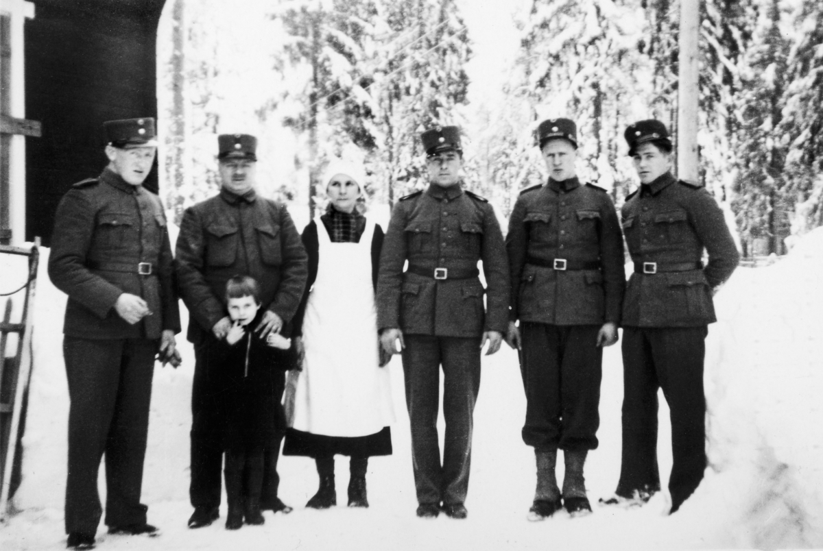 Gruppe 7 personer. 5 militære, 1 barn og en kvinne.
Bildet er tatt på Gardermoen våren 1939. 
Fra venstre: Nordli (fra Hamar), ukjent navn - men var sykkelreparatør fra Solør, har sitt barn foran seg, og sin kone ved siden av seg, hun var kokke. Håkon Holen, Adolfsen, Øivind Furulund fra Furnes. Alle de fem mennene er inne til 1. gangs militærtjeneste.