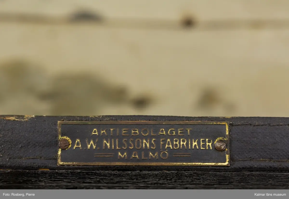 KLM 45797. Barnvagn, liggvagn. Stomme och hjul av svartlackerad metall, på stommen blå dekorlinjer. Gummiklädda hjul. Löstagbar vagnskorg med stomme av svartmålat trä med ljusblå dekorlinjer och fält målade i mörkare blå. På insidan är korgen klädd med vit galon. Vid korgens båda kortsidor handtag av vitt porslin. På vagnskorgens utsida ett vävt tygband av ylle i brun nyans. På ena kortsidan en liten metallskylt: AKTIEBOLAGET A.W. NILSSONS FABRIKER MALMÖ. Vagnen har en sufflett av svart galon med nedre kant av blått ylletyg. Upptill ett vävt tygband i gul nyans, på sidan ett handtag av vitt porslin. Vagnens ena fäste för suffletten är trasig och suffletten går därför inte att fästa ordentligt.