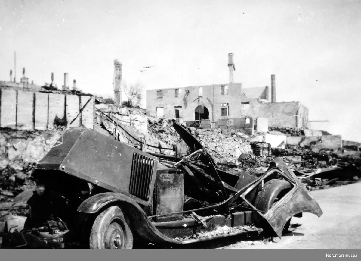 Bilen er en Renault fra 1920-tallet, med radiatoren montert bak motoren.(Info: Ivar Stav). Bilen er nyere enn 1924 da Renault fikk det rombiske emblemet ! (info:
Andreas Uddling)  - -Andre verdenskrig har kommet til Kirkelandet i Kristiansund. Her ser vi fra ruinene av Clausengården fra Storkaia, med et bilvrak i forgrunnen. Meieripipa bak th. Bildet er datert 28. april 1940, som er samme dag som byen ble bombet for første gang. serie. Fra Nordmøre museums fotosamlinger.