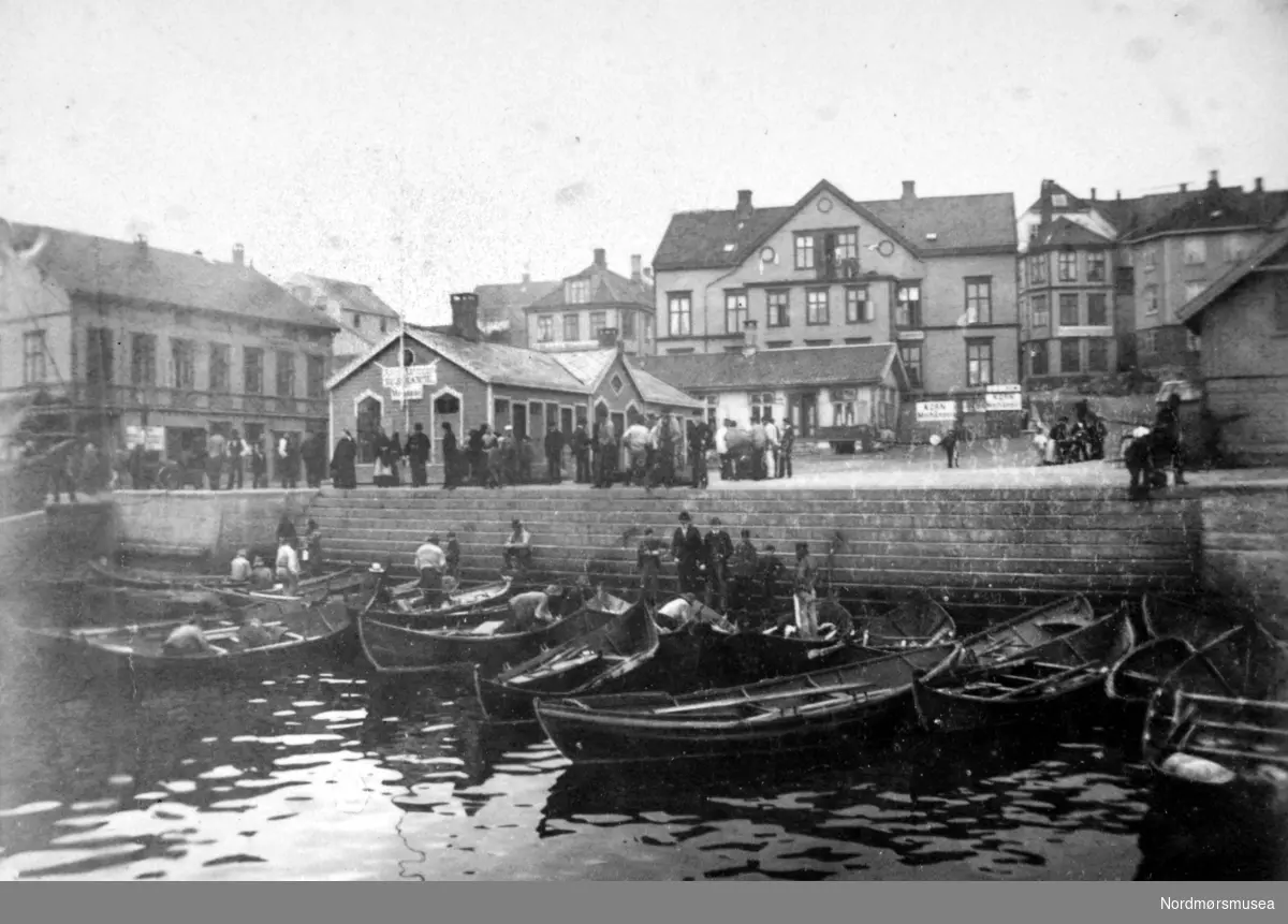 Et bilde fra Kirkeallmenningen, fisketorget og fisketrappa på Kirkelandet i Kristiansund, med atskillige småbåter, geitbåter og lystinger. I bakgrunnen ser vi bygningene rundt Kirkeallmenningen. Fra Nordmøre Museums fotosamlinger.